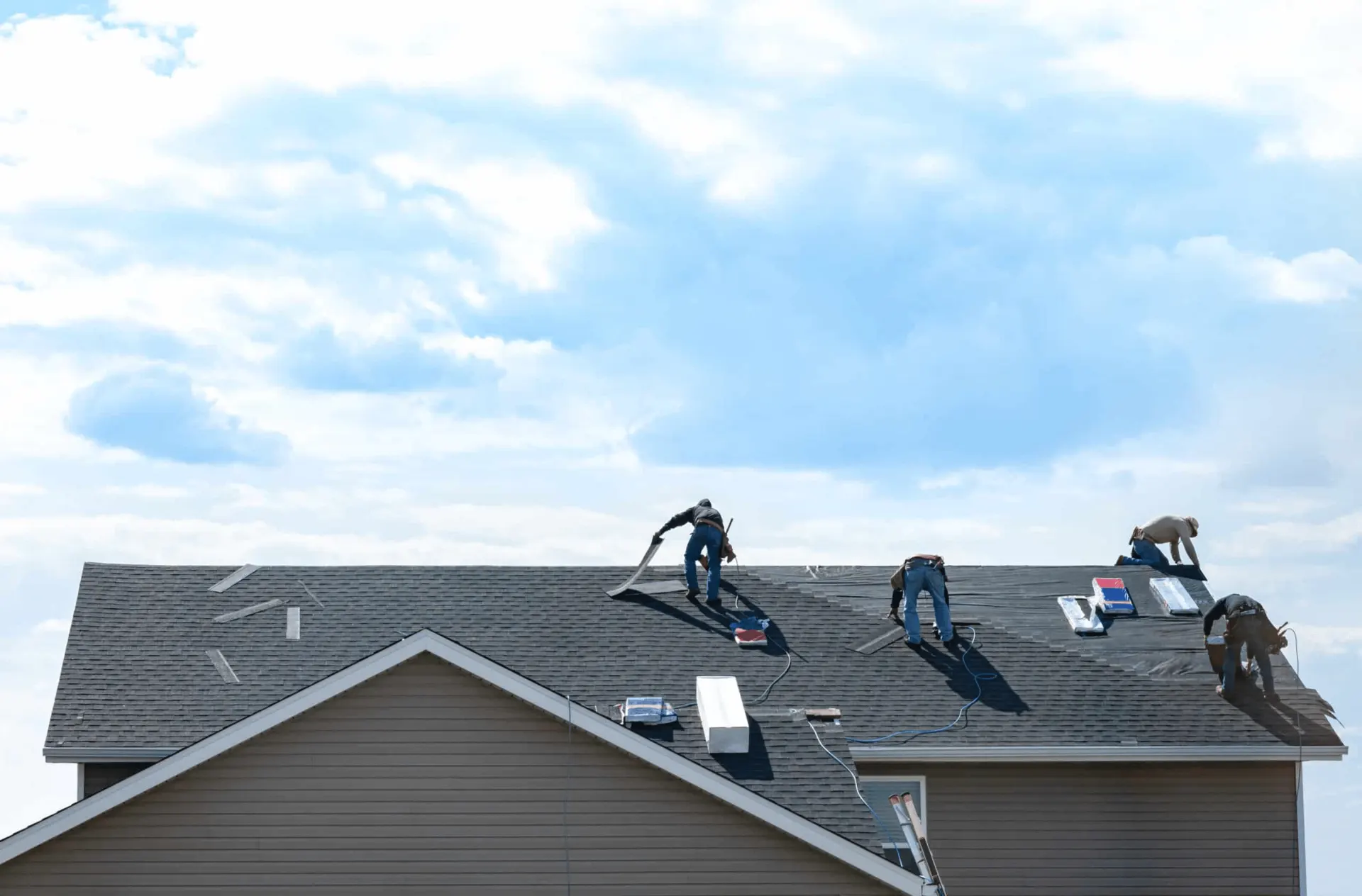 Multiple roofers are working to replace asphalt shingles on the roof of a large residential or commercial building under a bright, cloudy sky. Edmond OK Commercial Roof Replacement by Roof One.